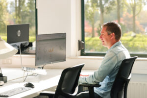 Man werkt achter zijn bureau met een dashboard vol grafieken op het scherm, in een zonnig kantoor met uitzicht op groen. De foto straalt focus en inzicht in personeelsplanning uit.