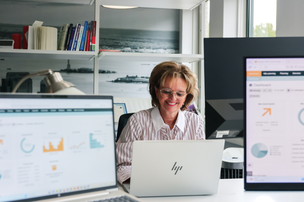 Vrouw werkt glimlachend achter haar laptop, omringd door schermen met grafieken en dashboards. De afbeelding straalt concentratie en plezier in datagedreven personeelsplanning uit.