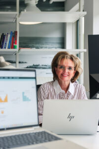 Vrouw met bril glimlacht achter haar laptop in een lichte kantoorruimte. Op de voorgrond zijn grafieken op andere schermen zichtbaar, wat samenwerking en datagedreven personeelsplanning benadrukt.