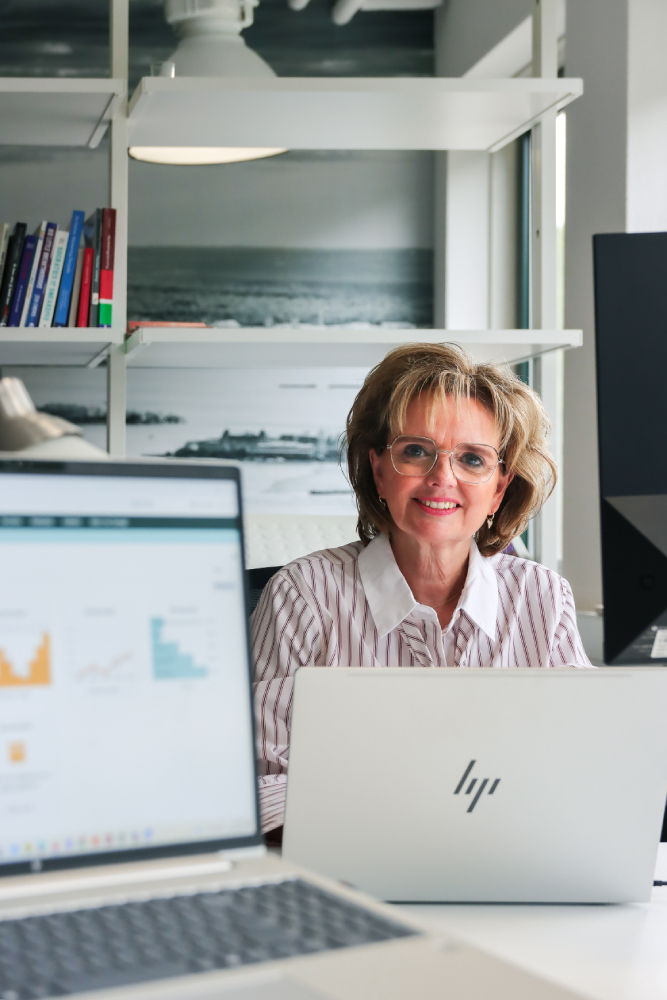 Vrouw met bril glimlacht achter haar laptop in een lichte kantoorruimte. Op de voorgrond zijn grafieken op andere schermen zichtbaar, wat samenwerking en datagedreven personeelsplanning benadrukt.