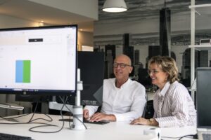 Twee collega’s zitten samen achter een computerscherm en bekijken een grafiek in een kantooromgeving. Ze overleggen geconcentreerd, wat samenwerking en inzicht in personeelsplanning weerspiegelt.