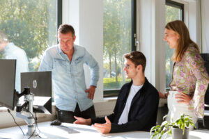 Drie collega’s overleggen bij een bureau met een computerscherm, terwijl zonlicht door de ramen valt. Ze bespreken samen een planningsscherm, symbool voor samenwerking en inzicht in personeelsplanning.