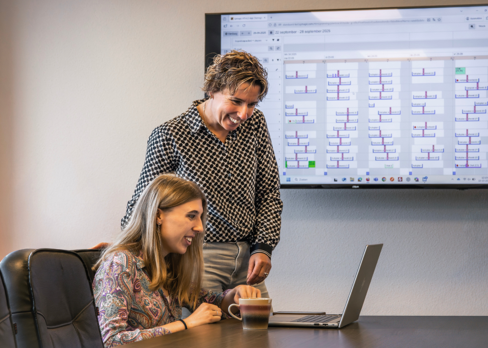 Twee collega’s bespreken samen een planning op een laptop, met op de achtergrond een groot scherm vol roosters. De sfeer is betrokken en professioneel, gericht op efficiënte personeelsplanning.
